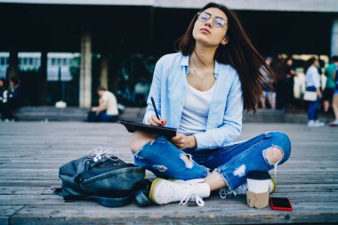 Şık giyinmiş, şık bir tasarım çizimi olan çekici bir genç kadın şehir ortamında oturmuş kompozisyon yazan hippi kız dışarıda ödev yapıyor ve Lotus pozisyonunda oturuyor.