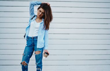 Sıradan bir kot giyip kameraya poz veren çekici bir genç kadının portresi şık kıyafetler giymiş, elinde kahve tutan çekici hippi kız promosyon arka planının yanında duruyor.