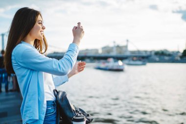 Gündelik kıyafetlerle gezen, gülümseyen ve blog için modern akıllı telefondan fotoğraf çeken çekici genç bir kadın. Hücresel hücrede güzel nehrin videosunu çeken hippi kız.