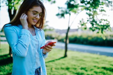 Modern akıllı telefon cihazında müzik uygulamasından kulaklıkla müzik dinleyen gülümseyen genç bir kadın parkın içinde duruyor. Pozitif hippi kız kentsel ortamdaki dijital cep telefonunda müzik listesinin keyfini çıkarıyor.