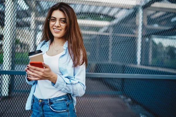 Portrait of beautiful young woman in stylish eyeglasses smiling at camera while standing in urban setting with coffee to go and modern mobile phone.Charming and positive hipster girl with smartphone