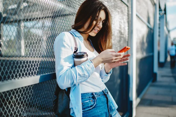 スタイリッシュな服を着たヒップスターブロガーは 都市部の設定に立って 無料の4Gインターネットを使用して現代のスマートフォン上のソーシャルネットワークでオンラインでチャットに行くためにコーヒーと — ストック写真