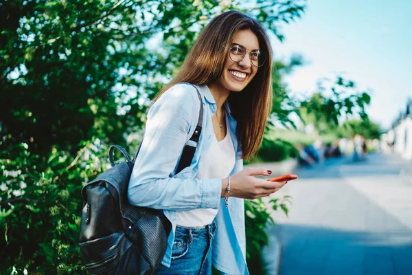 都市部の設定で笑ってバックパックと陽気な若い女性は手で現代のスマートフォンを保持しています パブリック4Gインターネット接続を使用してデバイス上のカジュアルウェア更新アプリで身に着けている積極的なヒップスターの女の子 — ストック写真