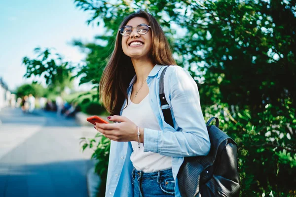 一个积极的嬉皮士女孩 背着背包在绿地里玩得很开心 快乐的年轻的高加索女人拿着数字手机站在城市里 天气很好 心情也很好 — 图库照片