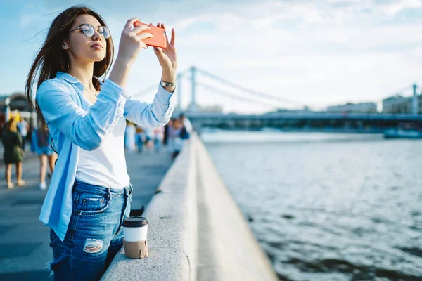 Charming young woman focusing camera on smartphone to make pictures for travel blog during summer trip in modern city.Attractive hipster girl shooting video for social networks in cellular