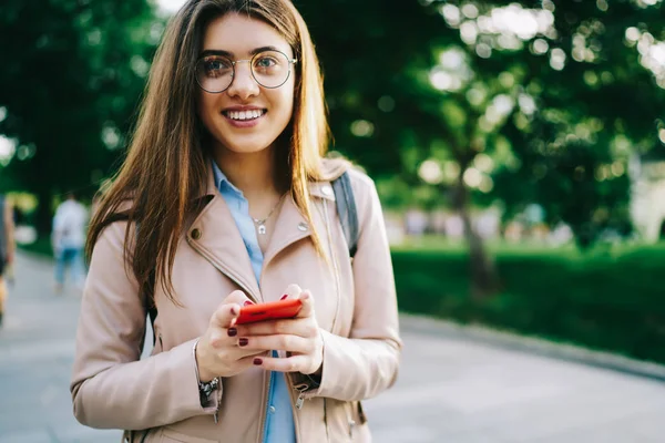 Portrait of cheerful young woman dressed in casual wear smiling at camera while typing text message on modern smartphone standing in urban setting.Positive hipster blogger updating profile on cellular