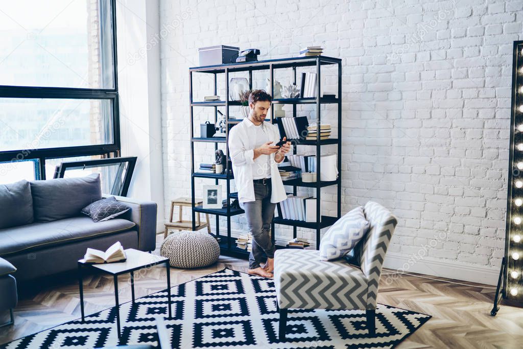Serious caucasian handsome man in casual wear standing in modern designed living room in apartment with mobile phone, hipster guy sending text messages on smartphone using app and 4G connection