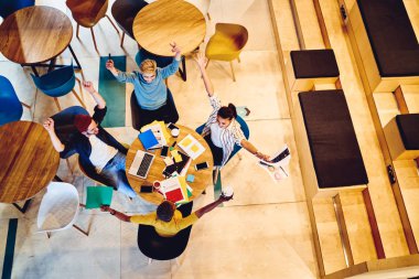 Mutlu hipster öğrencilerinin el ele tutuşup, kafenin içindeki projeyi tamamlamalarını kutluyorlar. Başarıyı kutlayan pozitif gençler eskizler ve dizüstü bilgisayarlarla aynı masada oturuyorlar.