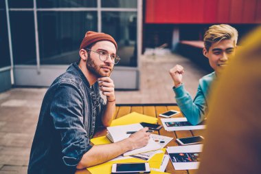 Öğrencilerle beyin fırtınası yaparken yaratıcı fikirler üzerine düşünerek gündelik giysiler giymiş düşünceli beyaz bir genç. Açık havada iş arkadaşlarıyla toplantı masasında oturan hippi çocuk.