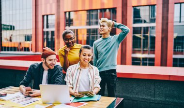 Gündelik giyinmiş çeşitli gençler modern akıllı telefondan komik şeyler izleyip dışarıda işbirliği yaparken dizüstü bilgisayarlarıyla oturuyorlar. 4G internet kullanan çok kültürlü hipster çocuklar.