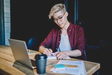 Gözlüklü zeki Asyalı genç adam dizüstü bilgisayarda webinar izlerken metin bilgileri yazıyor. Japon hippi öğrenci modern çalışma alanında dijital bilgisayarın başında ödev yapıyor.