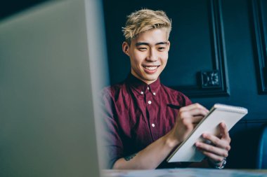 Modern dizüstü bilgisayarda Webinar 'ı izlerken notlar alan neşeli Çinli genç adam. Modern uzayda oturan notlara resim çizen pozitif Japon hipster öğrenci.