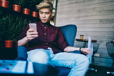 Modern akıllı telefondan 4G internete bağlı takipçilerden gelen tebligatı okuyan düşünceli Çinli hipster blogcusu modern uzay sandalyesinde oturuyor. Japon genç adam cep telefonuyla video izliyor.