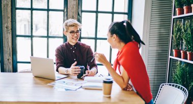 Modern ofiste tatilde birbirleriyle iletişim kuran neşeli Çinli öğrenciler, ahşap masada dizüstü bilgisayarla oturuyorlar. Pozitif Asyalı tasarımcılar arkadaşça sohbetin keyfini çıkarıyorlar.