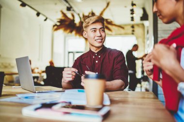 Sırıtkan Çinli hippi öğrenci dizüstü bilgisayarla masaüstünde oturuyor ve modern uzayda bayan meslektaşlarıyla birlikte iş kurma projesinde işbirliği yapma planını tartışıyor.