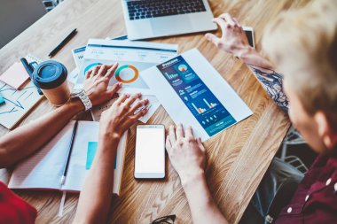 Masaüstünde kağıt diyagramları ile çalışan ve bilgi toplayan öğrencilerin en iyi kırpılmış görünümü. İstatistik çizelge üzerinde çalışan genç erkek ve kadın boş ekranlı akıllı telefonlu masada oturuyor.