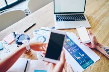 İnternet siteniz için fotokopi alanı olan modern akıllı telefonları tutan genç adam ve kadının kırpılmış görüntüsü veya ahşap masaüstünde dizüstü bilgisayarlı ve diyagramlı kağıt çizimleriyle oturan içerik