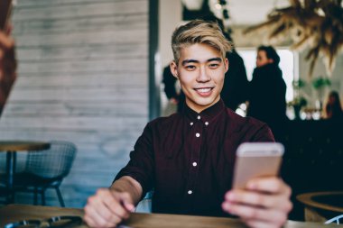 Neşeli Çinli yenilikçi adam sosyal ağlarda akıllı telefondan komik videolar izliyor modern mekanda oturuyor. Japon blogcu kendi web sitesinden 4G internet kullanarak yayın indiriyor.