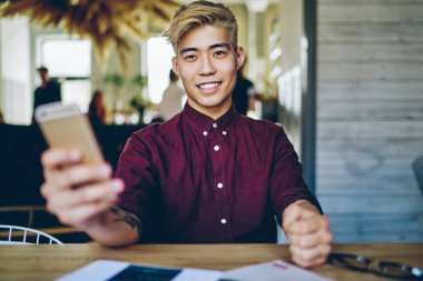 4G interneti kullanarak akıllı telefon cihazının profilini güncellerken gülümseyen Çinli pozitif hipsterin portresi. Masaüstünde oturan yakışıklı Asyalı öğrenci cep telefonu tutuyor.