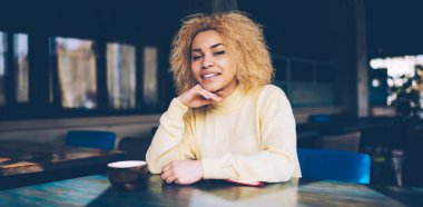 En sevdiği kafeteryadaki boş zamanlarında kameraya gülümseyen sıradan giyinmiş mutlu koyu tenli hippi kız portresi. Kafede sarı kıvırcık saçlı Afro-Amerikan kadın.