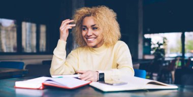 Koyu renk tenli, günlük kıyafetler giymiş, kafeterya masasında oturmuş üniversite projesinde ders kitabı okuyan, kıvırcık saçlı mutlu siyah hippi kız.