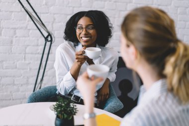 Sevimli, koyu tenli genç bir kadın elinde bir fincan kahve tutuyor ve modern dairede buluşurken en iyi arkadaşıyla iletişim kuruyor. Oldukça Afrikalı Amerikalı hippi kız iş arkadaşıyla gülüyor.