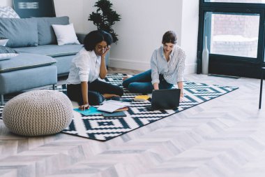 Kalifiye Afro-Amerikan ve Kafkasyalı öğrenciler yerde oturup laptop bilgisayar ve kablosuz internet kullanarak modern stil apartmanda ortak yayın üzerinde işbirliği yapıyorlar.