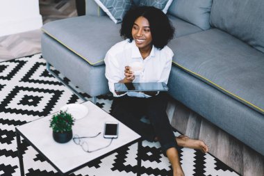 Kahvesini ve dijital dokunmatik defterini elinde tutarak gülen koyu tenli hippi blogcu modern bir dairede şık bir iç dekorasyonla oturuyor. Neşeli, düşünceli, Afro-Amerikan genç kadın.