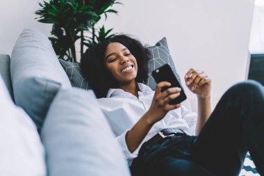 Pozitif Afro-Amerikan genç kadın rahat koltukta dinleniyor ve akıllı telefondan komik haberler okuyor evde boş zamanların tadını çıkarıyor. Neşeli blogcu, sosyal ağlarda çok beğeni toplayan telefonla konuşuyor.