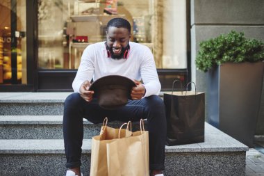 Alışveriş saatinden sonra merdivenlerde dinlenen, şık şapkalara bakan ve bir şeyler satın aldığı için kendini iyi hisseden, kağıt torbalı mutlu hippi erkek.