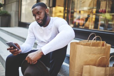 AVM 'nin yanındaki merdivenlerde oturan Afro-Amerikalı adamın portresi, alışveriş sonrası elinde akıllı telefon cihazı olan esmer tenli bir erkek alışveriş meraklısı dinleniyor.