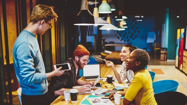 Happy crew of trendy dressed group of hipsters discussing ideas for startup sitting at meeting table with netbook and stationery, multiracial crew of students having brainstorming session togethe