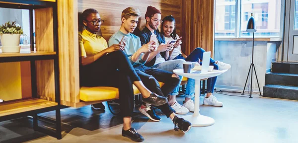年轻的时髦男女嬉皮士们一起坐在合作的空间里 使用着沉迷于互联网的现代手机 年轻的多种族女性和男人通过利索尔上的智能手机结成网络 — 图库照片