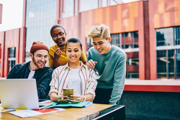 Cheerful diverse young people dressed in casual wear laughing while watching funny videos in blog on modern smartphone using 4G internet connection outdoors.Positive diverse hipsters installing app