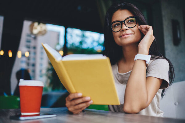 Dreamy caucasian young woman thinking about romantic novel spending free time on literature in cafe, positive hipster girl in spectacles for vision correction looking away enjoy interesting book