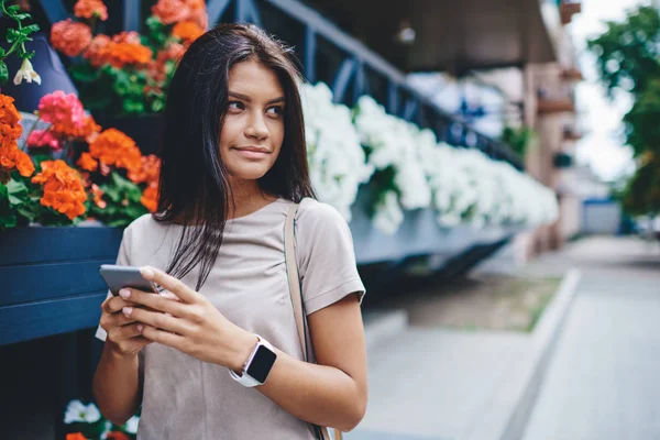 Beautiful brunette female looking away standing on street near flower beds with smartphone connected to 4G, caucasian woman looking away using modern smartphone for networking outdoors in spring