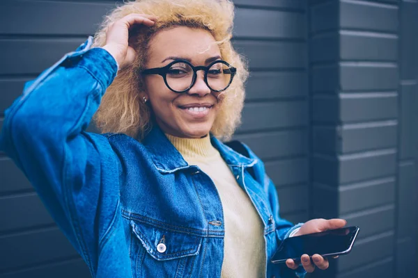 Close up portrait of cheerful Afro American hipster girl in eyewear for provide eyes protection holding smartphone gadget for browsing internet in hand and smiling at camera on urban setting