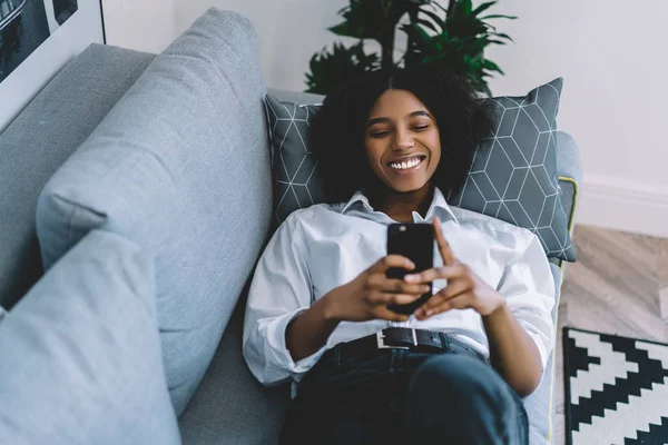 在家里用4G网络在智能手机上边聊天边笑边笑的快乐的嬉皮士博主 积极的非洲裔美国女青年在沙发上放松 并与朋友聊天 — 图库照片