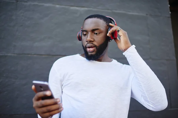 Male meloman searching new audio songs on content website feeling shocked from application price, man chatting online via mobile phone during time for listening audio book via bluetooth headphones