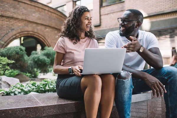 Happy male and female friends from America or Africa enjoying live communication and time for discussing audio book from laptop device, cheerful dark skinned couple in love talking on urban setting