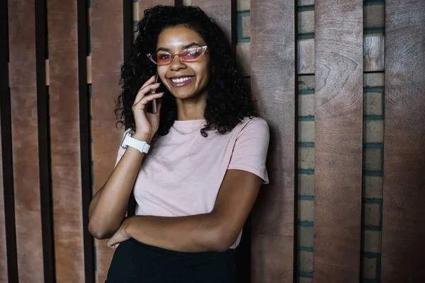 Half length portrait of excited female teenager dressed in trendy apparel posing on urban setting while making friendly mobile conversation via cellphone application, cheerful woman in pink sunglasses