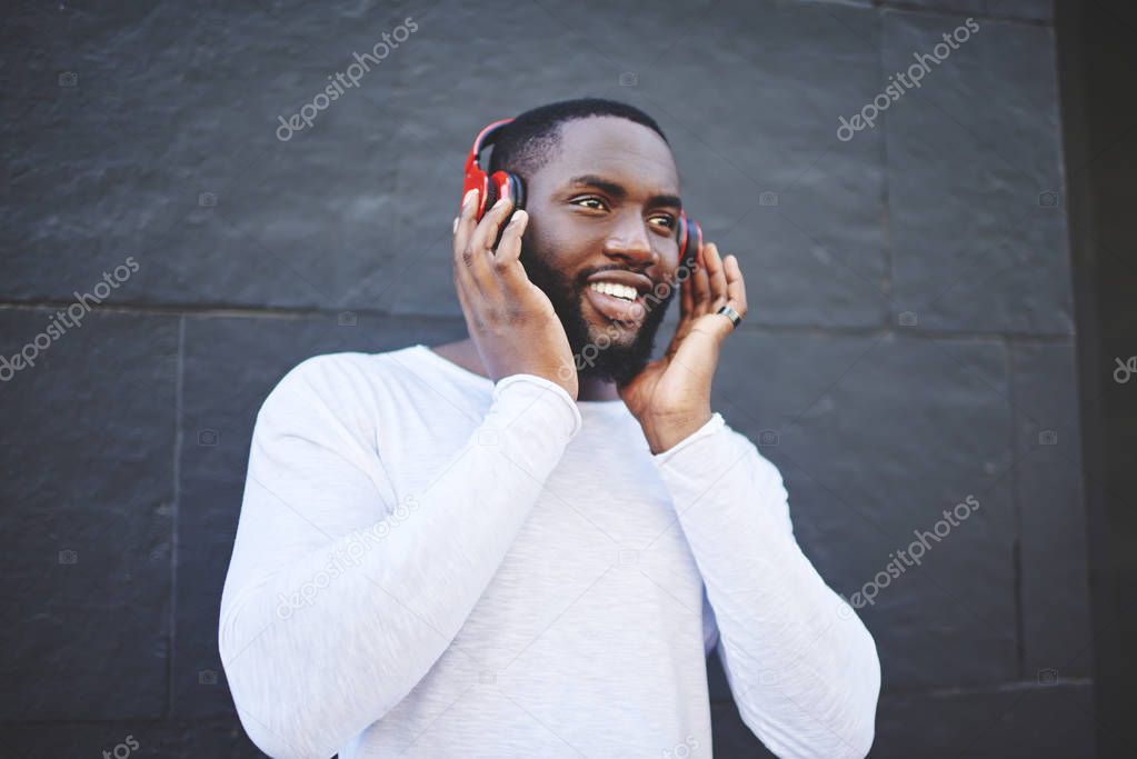 Carefree male meloman listening music with high volume using headphones at publicity area, positive smiling hipster guy enjoying favourite music playlist via earphones during leisure time outdoors