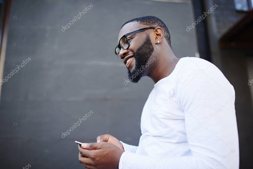Side view of cheerful male blogger in optical eyewear for vision correction looking away while waiting for email from friend, happy smiling hipster guy with cellphone enjoying time on publicity area