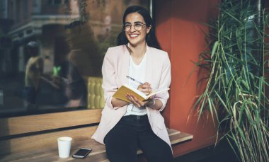 Gözlüklü mutlu yeniyetme kız göz korumasını sağladığı için gülümserken üniversite kompozisyonu hazırlıyor. Heyecanlı bir fikirle şehir merkezindeki kahve dükkanının yanında oturuyor. Notnotlu pozitif öğrenci.