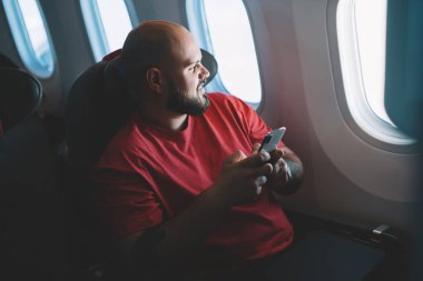 El ele tutuşan genç adam koltukta kendini rahat hissediyor. Uçağın kabin penceresinin yanında otururken internete bağlanan gülümseyen erkek uçuş yolcusu. Uçak kipi