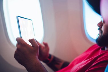Beyaz erkek uçak yolcusu, kabinde uçak penceresinin yanında otururken tablet ekrana dokunuyor, erkek, kablosuz internet bağlantısı ile uçuşun keyfini çıkarıyor.