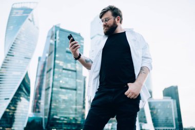 Gözlüklü beyaz adam cep telefonuyla sohbet okudu. 4G internete bağlandı. Arka planda gökdelenler var. Milenyum erkek yolcusu akıllı telefon kontrol rotasında navigasyon kullanıyor.