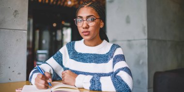 Üniversite kafeteryasında okul ödevi yapan konsantre kız öğrenci, moda gözlüklü akıllı düşünceli kadın portresi kameraya bakıyor ve sınava hazırlanıyor.