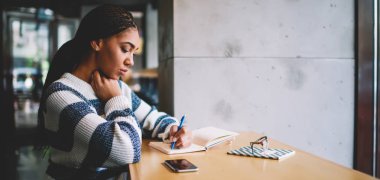 Düşünceli bayan öğrenci, kafeteryada oturmuş, boş zamanlarında not defterinde otokopi notları üzerine çalışan konsantre Afrikalı bir kadın, eğitim için ders kitabı yazıyor.