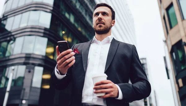 以下是一位穿着正装 手持手机和咖啡站在城市环境中的商人的照片 他们带着沉思的目光向远方看去 一位善于思考的金融专家带着智能手机站在城市环境中 — 图库照片